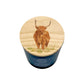 A scented candle in a dark blue cylindrical tin with a round wooden lid featuring an illustration of a Highland cow on a grassy field.