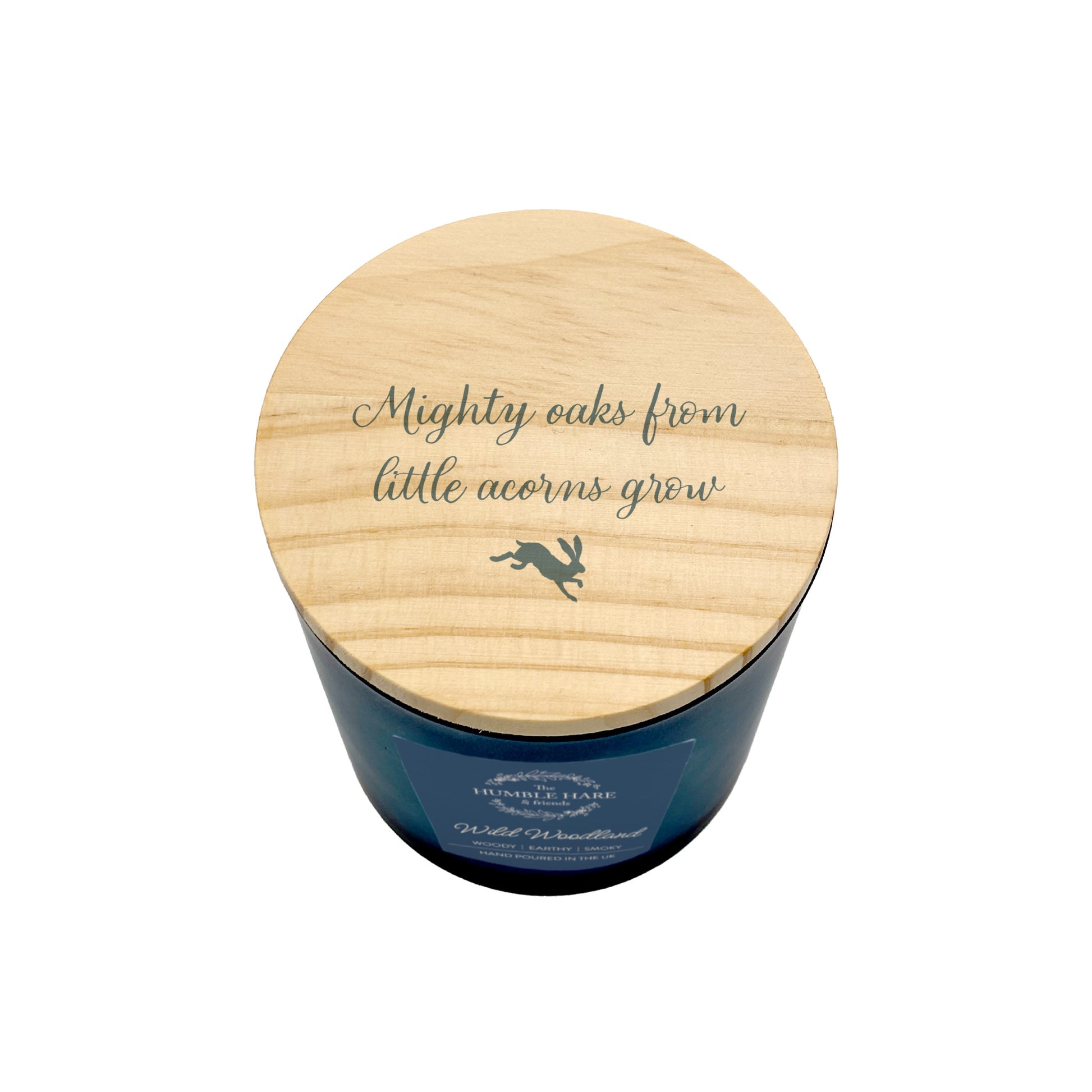 A blue glass jar candle with a light natural wood lid, featuring the quote 'Mighty oaks from little acorns grow' and a small leaping hare silhouette. 