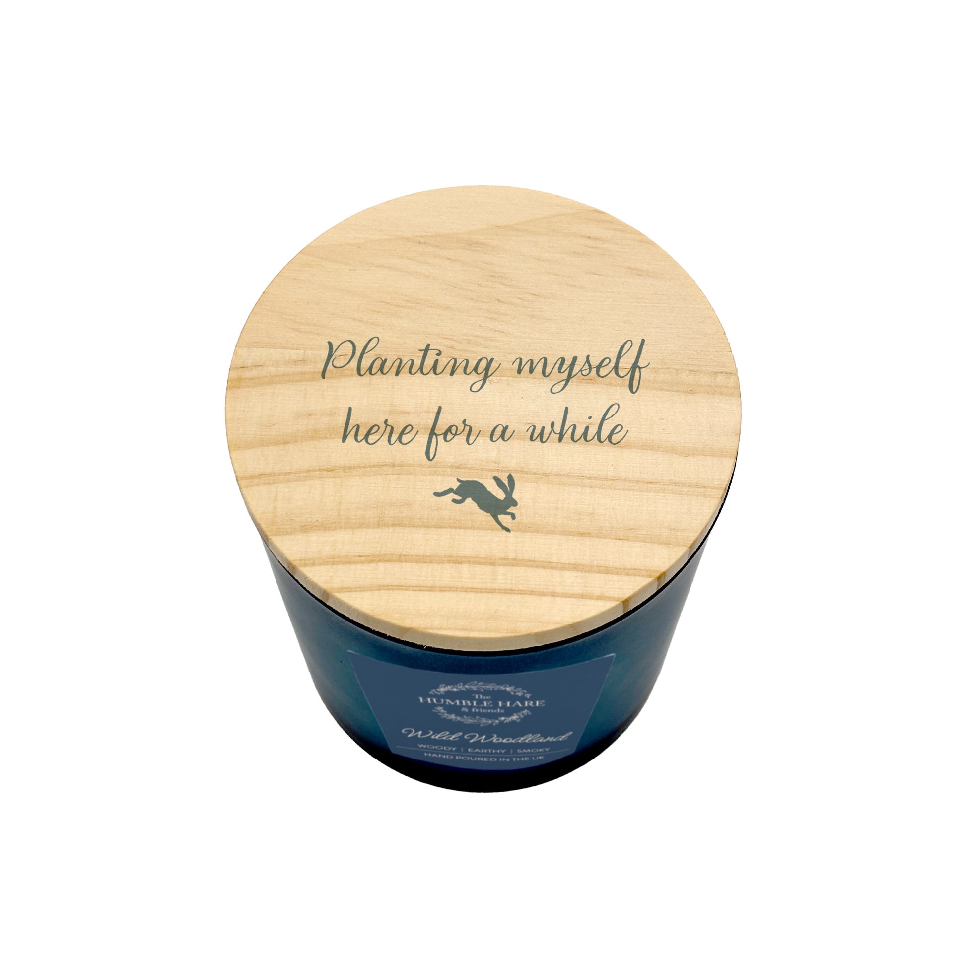A dark blue glass jar candle with a light natural wood lid with the phrase 'Planting myself here for a while' and a small hare silhouette, shown on a white background.