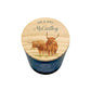 A personalised scented candle in a dark blue glass jar with a round wooden lid. The lid features an illustration of two Highland cows and is personalized with the text 'MR & MRS McCarthy'.