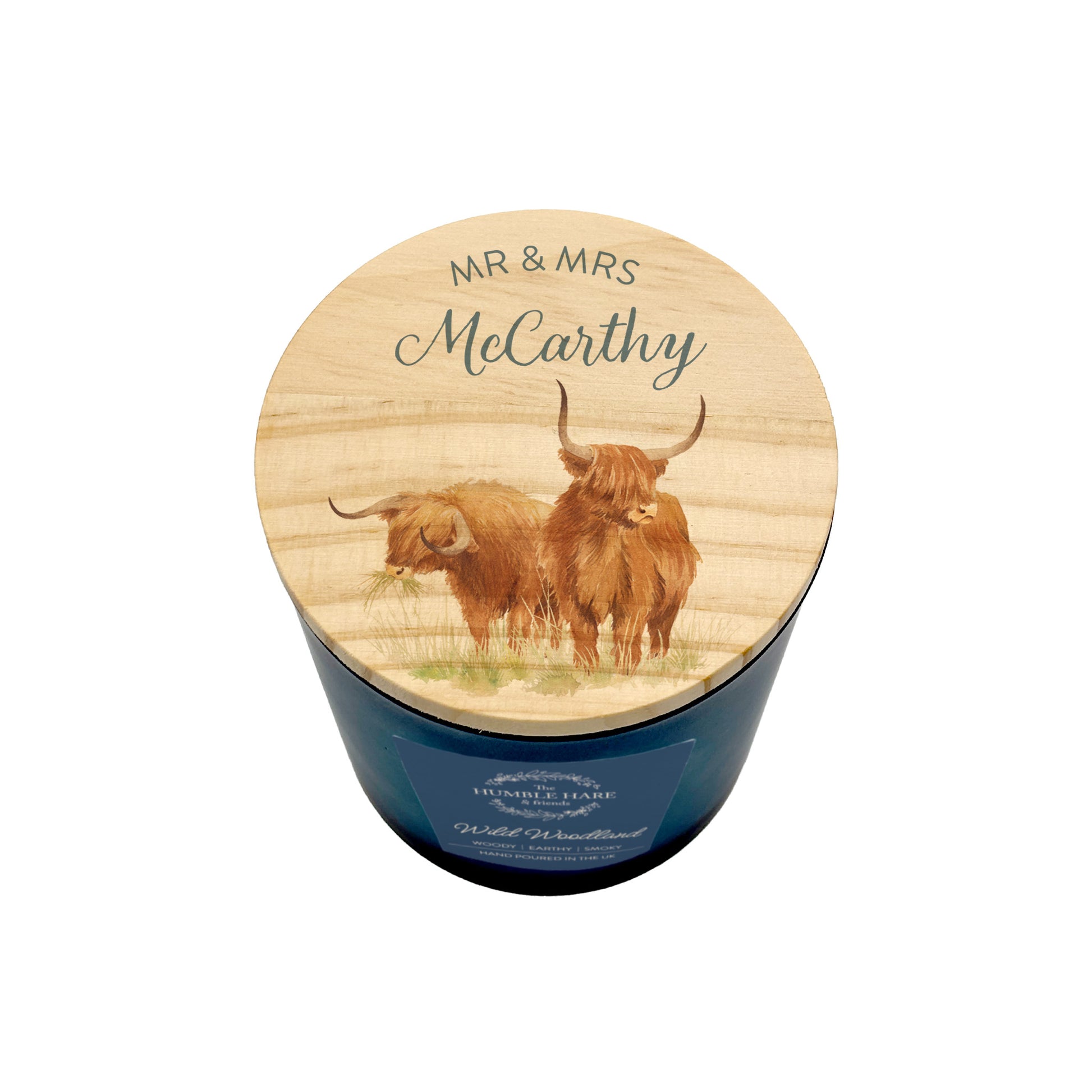 A personalised scented candle in a dark blue glass jar with a round wooden lid. The lid features an illustration of two Highland cows and is personalized with the text 'MR & MRS McCarthy'.