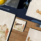 A square marble coaster with a design of a border collie working dog herding sheep through a gate on the farm. The coaster is set on a wooden country kitchen table.