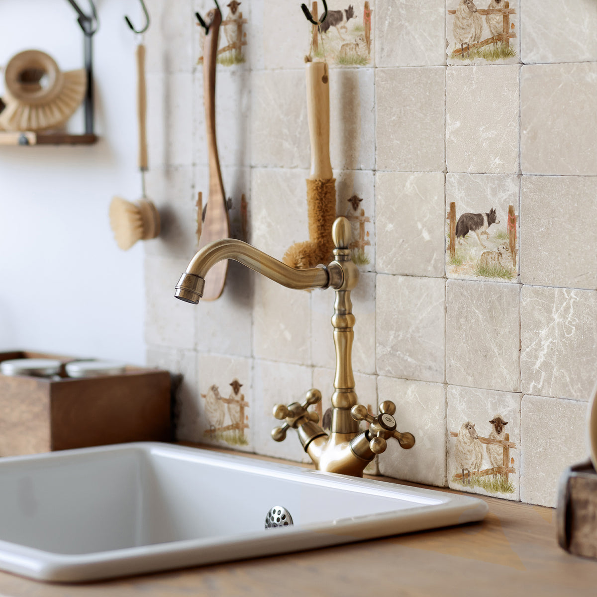 A close-up of a kitchen sink with a polished brass faucet mounted against a beige tiled backsplash featuring a border collie dog and sheep pattern. 