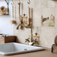 Rustic farmhouse kitchen sink with a vintage-style brass faucet, set against beige stone tiles decorated with border collie and sheep illustrations. Wooden utensils hang on hooks on the wall.