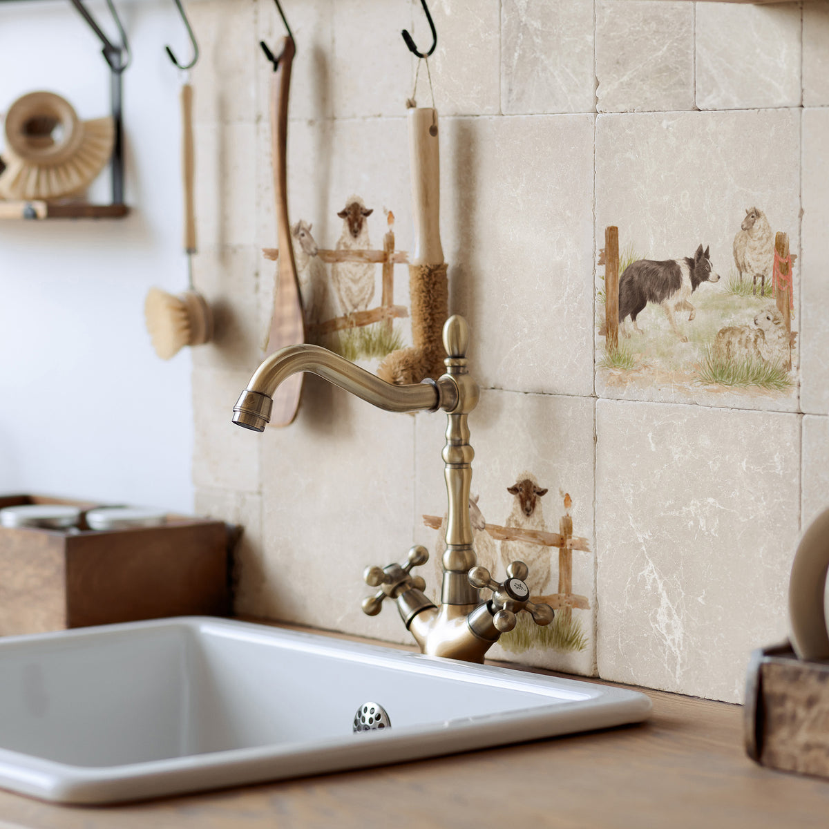 Rustic farmhouse kitchen sink with a vintage-style brass faucet, set against beige stone tiles decorated with border collie and sheep illustrations. Wooden utensils hang on hooks on the wall.