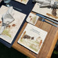 A square medium marble platter with a design of a border collie working dog herding sheep through a gate on the farm. The platter is personalised with custom text and set on a wooden country kitchen table.