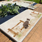 A rectangular marble sharing platter with a watercolour design of a border collie working dog herding sheep through a gate on the farm. The platter is set on a wooden country kitchen table.