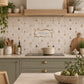 A styled kitchen interior with beige mosaic tile backsplash featuring a small bird and tree pattern, a central decorative sign reading 'Club Tree House', sage green cabinets, a black induction cooktop, and wooden shelving with plants and ceramic vessels.