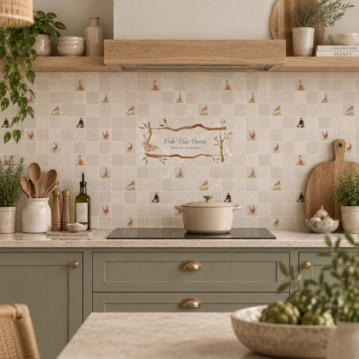 A styled kitchen interior with beige mosaic tile backsplash featuring a small bird and tree pattern, a central decorative sign reading 'Club Tree House', sage green cabinets, a black induction cooktop, and wooden shelving with plants and ceramic vessels.