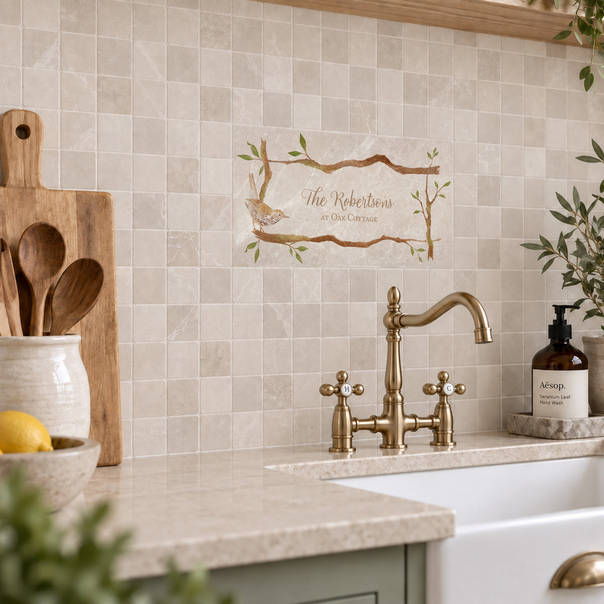 A kitchen backsplash made of light beige square mosaic tiles, featuring a custom printed decorative tile with a bird and the text 'The Roberts at One Corner' above a brass bridge faucet and white farmhouse sink.