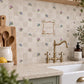 A styled kitchen interior with beige mosaic tile backsplash featuring a small bird and tree pattern, a central decorative sign reading 'Club Tree House', sage green cabinets, a black induction cooktop, and wooden shelving with plants and ceramic vessels.