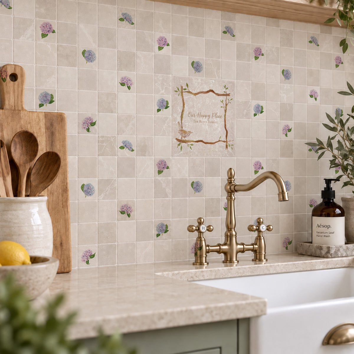 A styled kitchen interior with beige mosaic tile backsplash featuring a small bird and tree pattern, a central decorative sign reading 'Club Tree House', sage green cabinets, a black induction cooktop, and wooden shelving with plants and ceramic vessels.