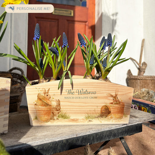 An arched wooden planter box full of spring bulbs. The planter box shows a watercolour design of cute bunny rabbits in plant pots, eating the flowers. The planter can be personalised with names and a selection of phrases to suit any occasion, including wedding, anniversary, engagement, birthday and new home.