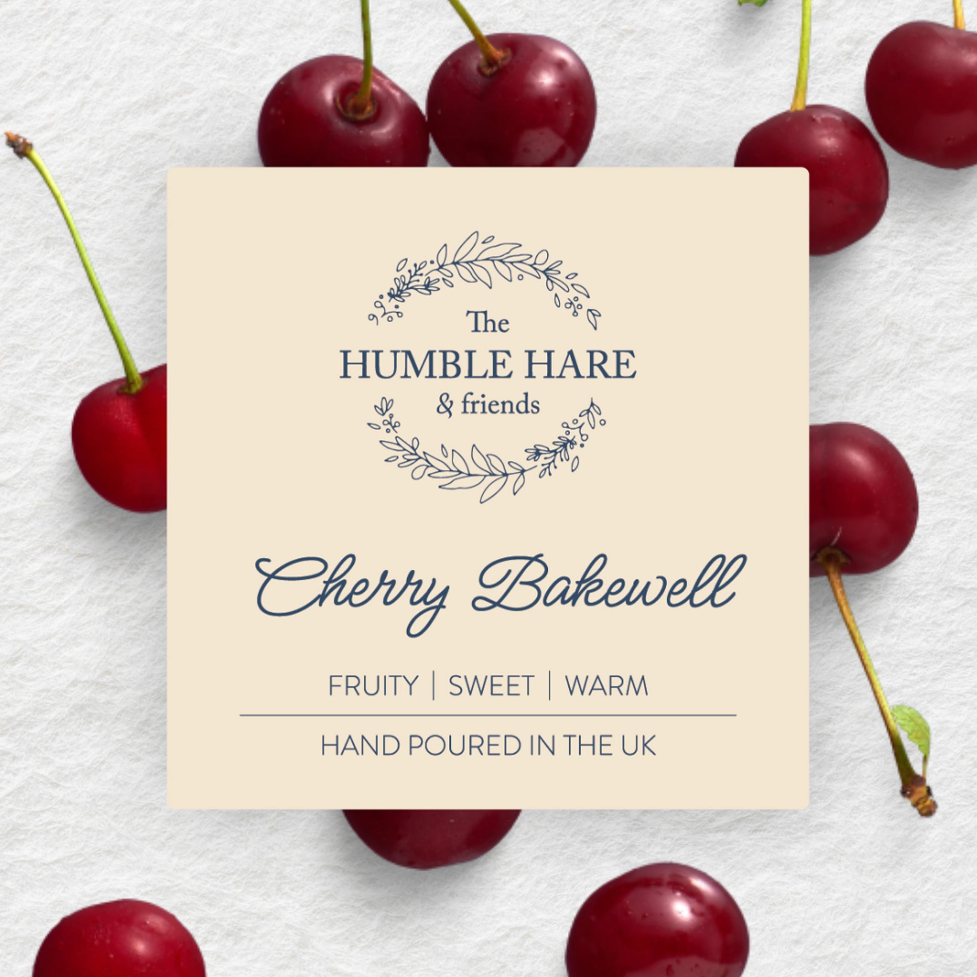 A square cream-colored label for The Humble Hare's Cherry Bakewell scented candle surrounded by fresh red cherries on a white textured background.