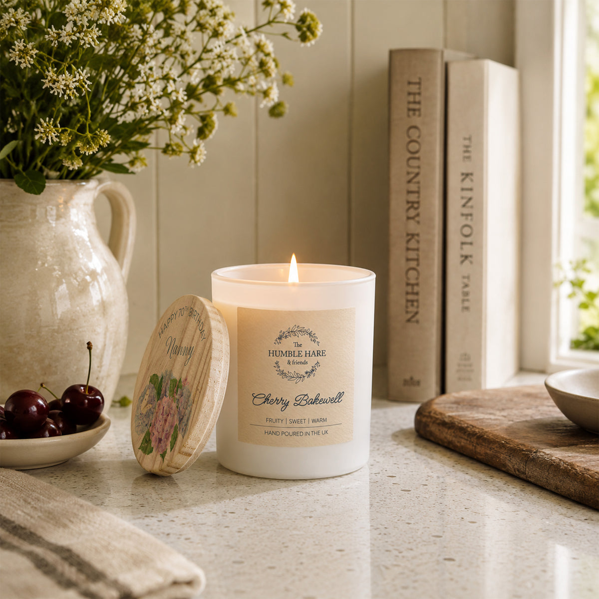 A lit white candle with a personalised wooden lid featuring a birthday message and hydrangea flowers sits on a kitchen counter beside a ceramic pitcher of flowers, cherries, books, and a cutting board.
