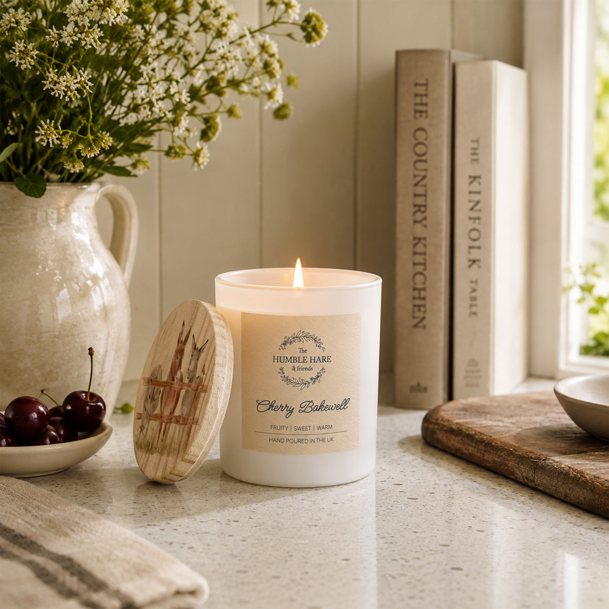 A lit white candle with a wooden lid featuring donkeys sits on a kitchen counter beside a ceramic pitcher of flowers, cherries, books, and a cutting board in a bright, styled interior.