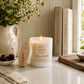 A lit white candle with a personalised wooden lid featuring a good luck bunny design sits on a kitchen counter beside a ceramic pitcher of flowers, cherries, books, and a cutting board in a bright, styled interior.