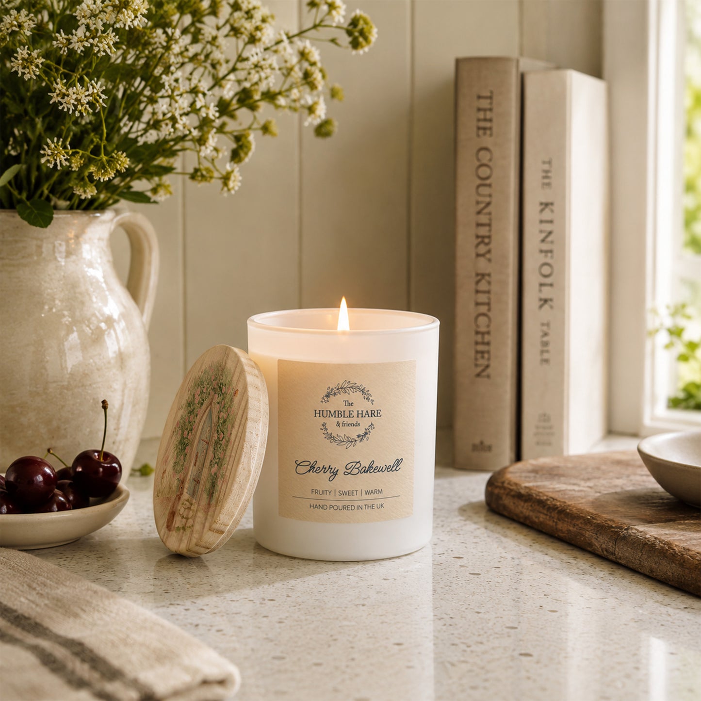 A lit white candle with a wooden lid featuring a watercolour hidden garden design sits on a kitchen counter beside a ceramic pitcher of flowers, cherries, books, and a cutting board in a bright, styled interior.