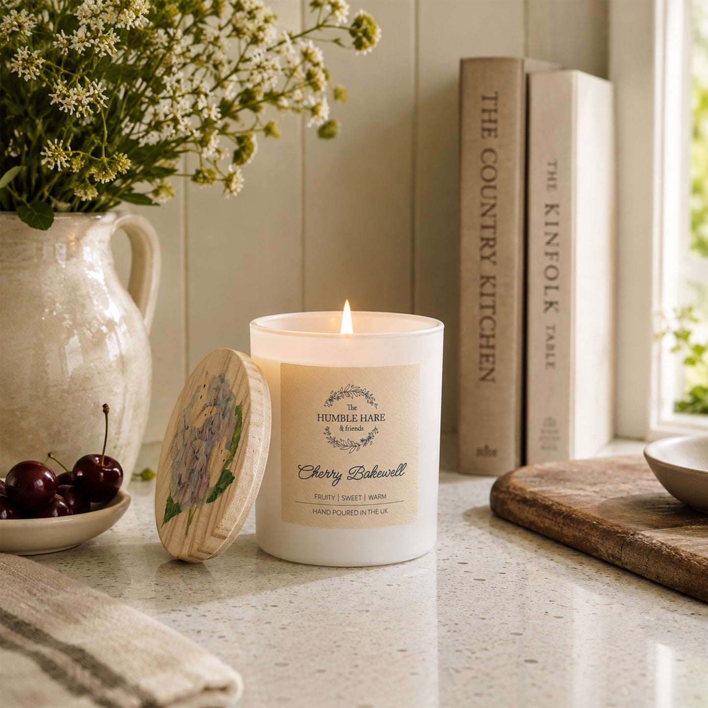 A lit white candle with a wooden lid featuring a blue hydrangea flower sits on a kitchen counter beside a ceramic pitcher of flowers, cherries, books, and a cutting board in a bright, styled interior.