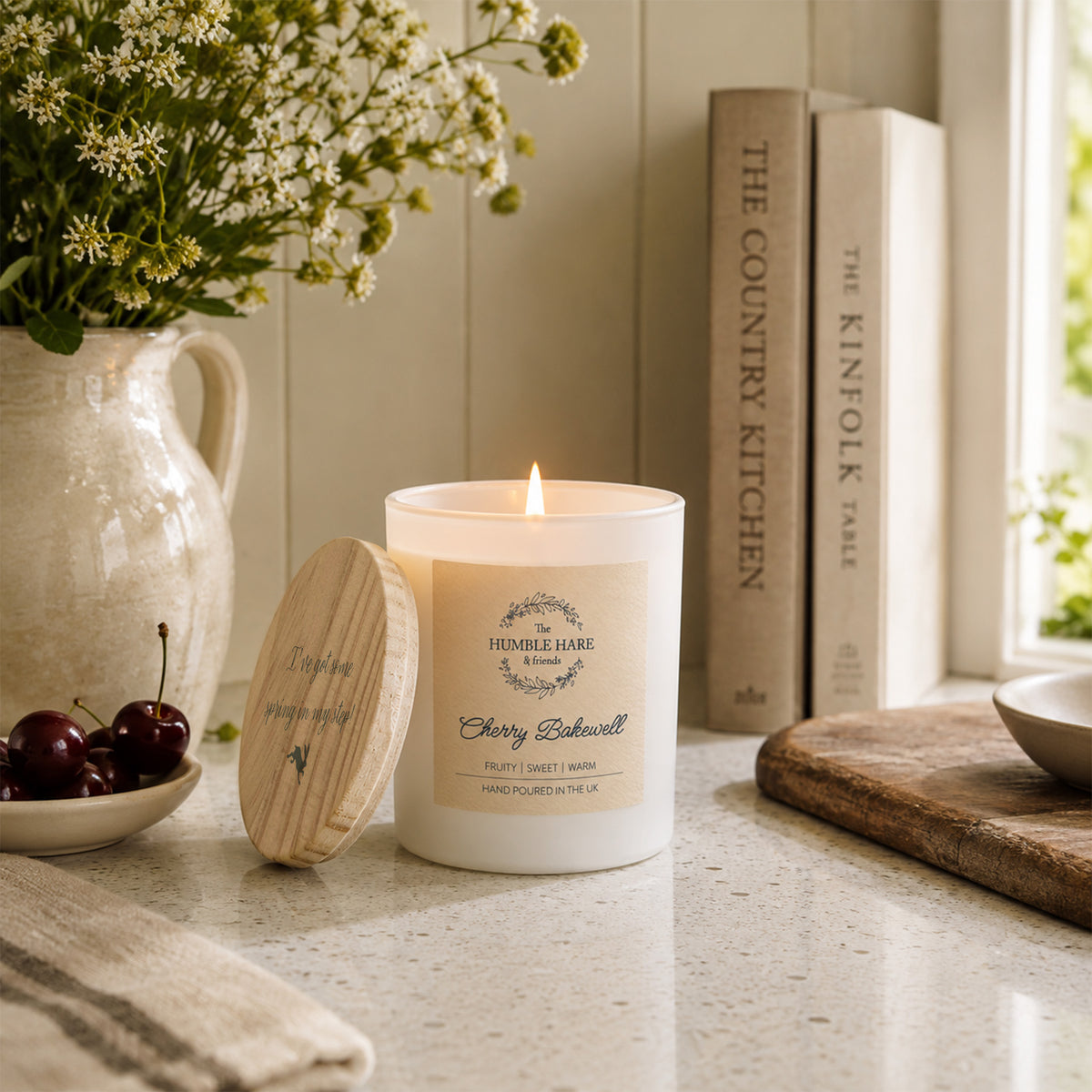 A lit white candle with a wooden lid featuring a quote sits on a kitchen counter beside a ceramic pitcher of flowers, cherries, books, and a cutting board in a bright, styled interior.