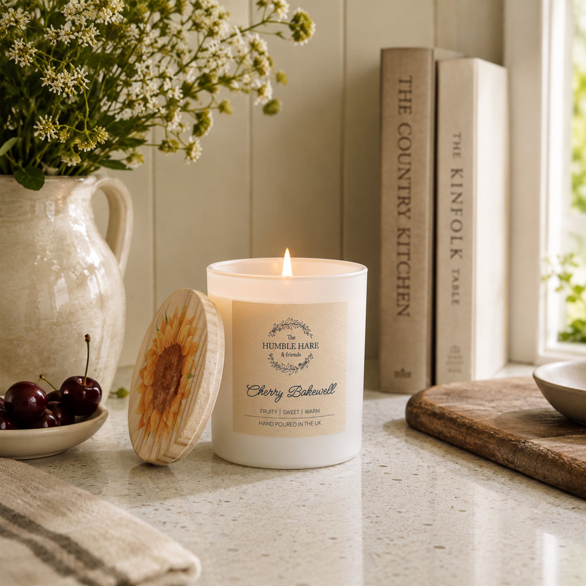 A lit white candle with a wooden lid featuring a sunflower sits on a kitchen counter beside a ceramic pitcher of flowers, cherries, books, and a cutting board in a bright, styled interior.