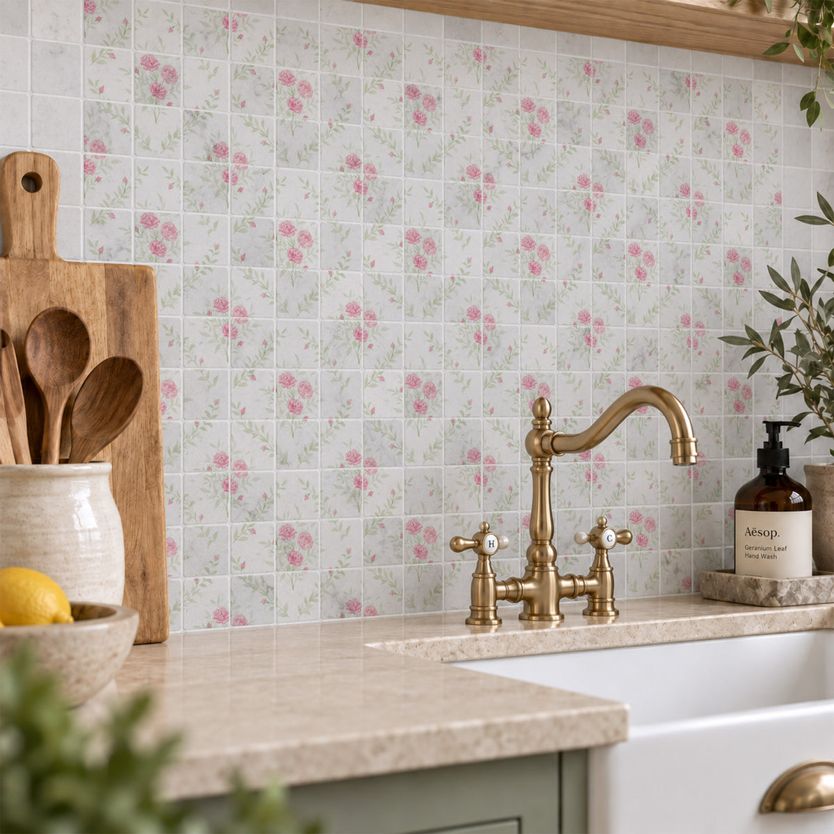 A kitchen backsplash made of small square tiles with a delicate pink floral pattern on a light background, installed behind a white farmhouse sink with a brass faucet. The scene includes a beige stone countertop, wooden utensils in a ceramic jar, and a soap dispenser.