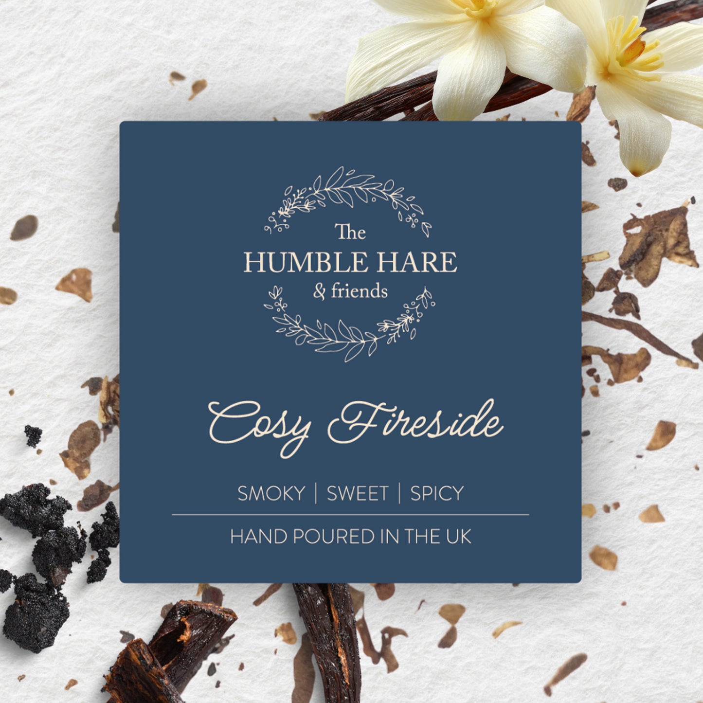 A square dark blue product label for The Humble Hare's Cosy Fireside vanilla and tobacco scented candle, styled on a white textured surface with vanilla flowers and pods.
