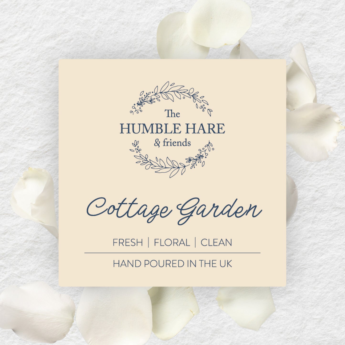 A square cream-colored label for 'The Humble Hare & friends' candle scent 'Cottage Garden', styled on a textured white background with scattered white rose petals.