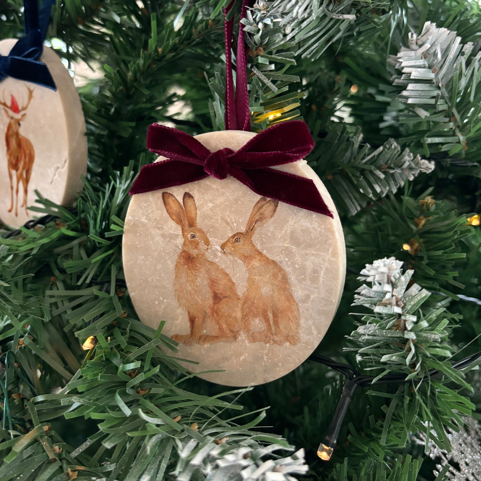 An oval marble bauble featuring a watercolour hare couple, with the option to add personalised text to celebrate the couple's first Christmas. The bauble is finished with a velvet ribbon in a choice of colours.