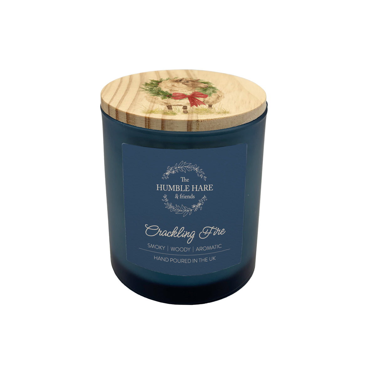 A dark blue glass jar candle with a light wood lid decorated with a sheep wearing a Christmas wreath illustration. The front features a blue label with white text that reads 'HUMBLE HARE & FRIENDS' and 'Crackling Fire'.