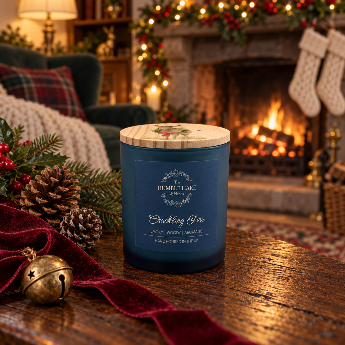 A lit blue glass candle jar labelled 'The Humble Hare & Friends Cracking Fire' and a wooden lid featuring a watercolour design of a donkey wearing a Christmas wreath. The Christmas candle sits on a speckled tray in a cosy holiday setting with pine branches, a fireplace, wrapped gifts, and a plaid blanket in the background.