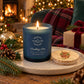 A lit blue glass candle jar labelled 'The Humble Hare & Friends Cracking Fire' and a wooden lid featuring a watercolour design of a Highland cow wearing a Christmas wreath. The Christmas candle sits on a speckled tray in a cosy holiday setting with pine branches, a fireplace, wrapped gifts, and a plaid blanket in the background.