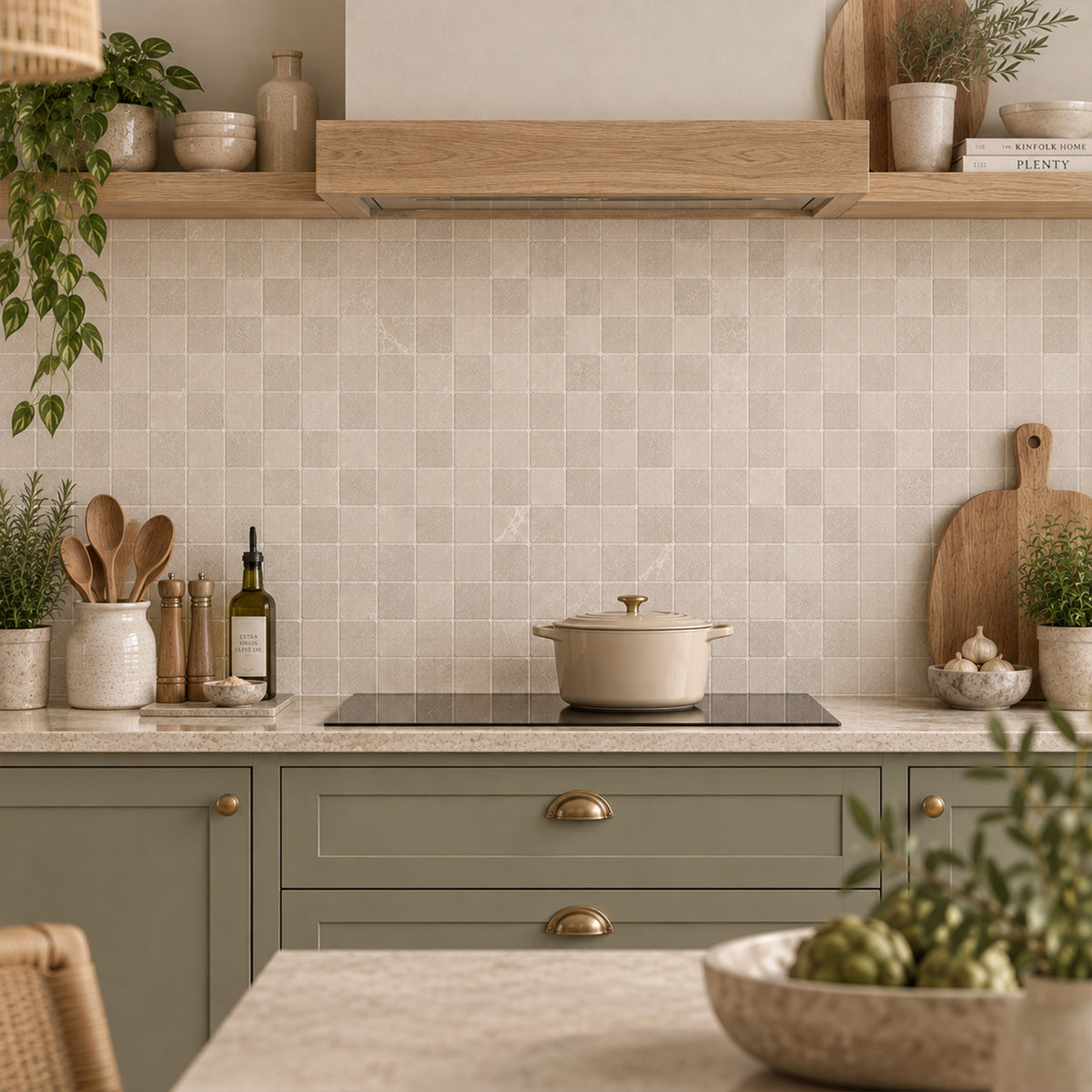 A rustic country kitchen with an induction hob and sage green cabinets in front of a wall tiled with plain cream marble mosaic tile sheets to create a splashback.