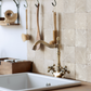 An antique brass gooseneck kitchen faucet with cross handles mounted on a cream marble tiled backsplash above a white sink, with hanging wooden kitchen tools in the background.