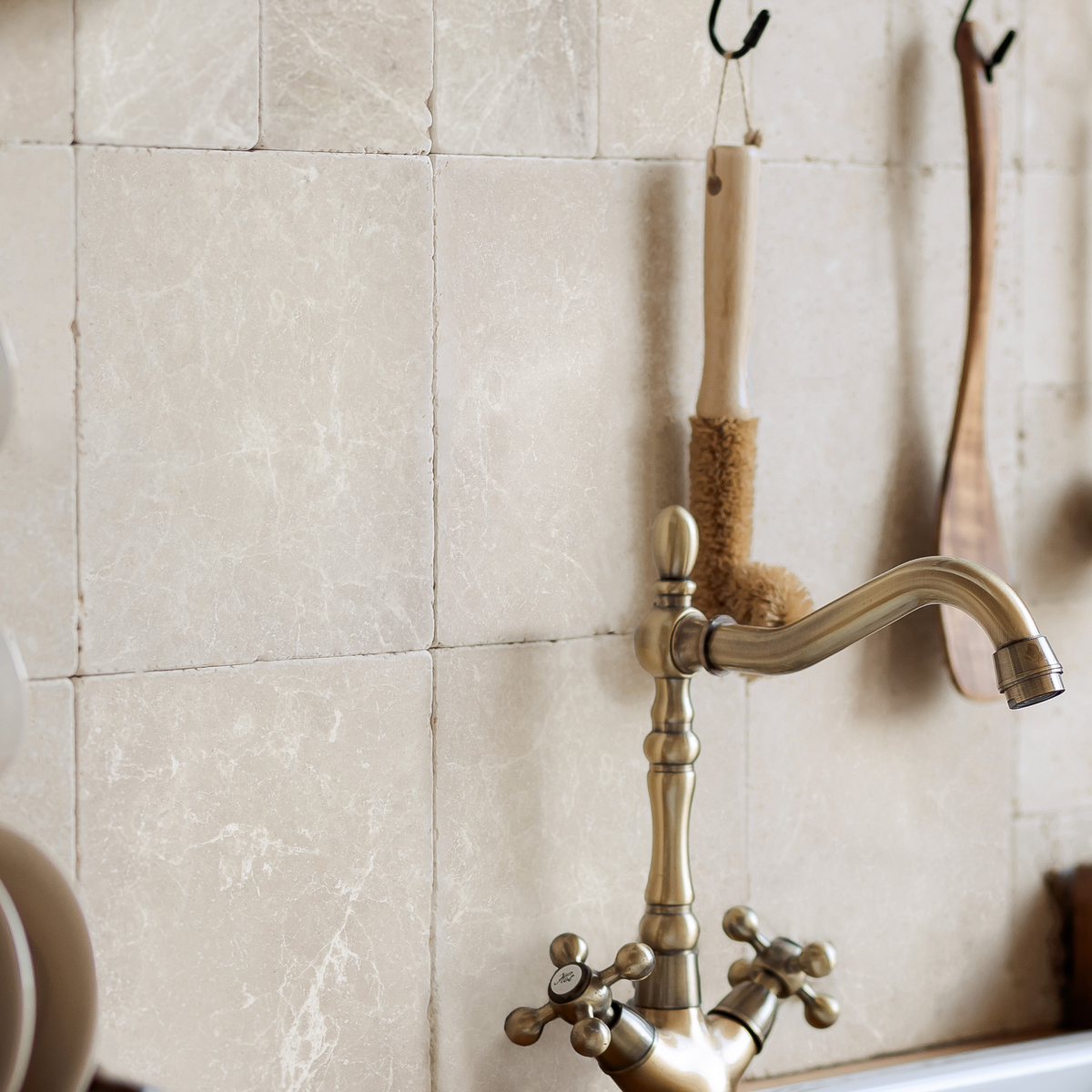 An antique brass kitchen faucet with cross handles mounted against a beige marble tile wall, with hanging scrubbers and a wooden spoon visible.
