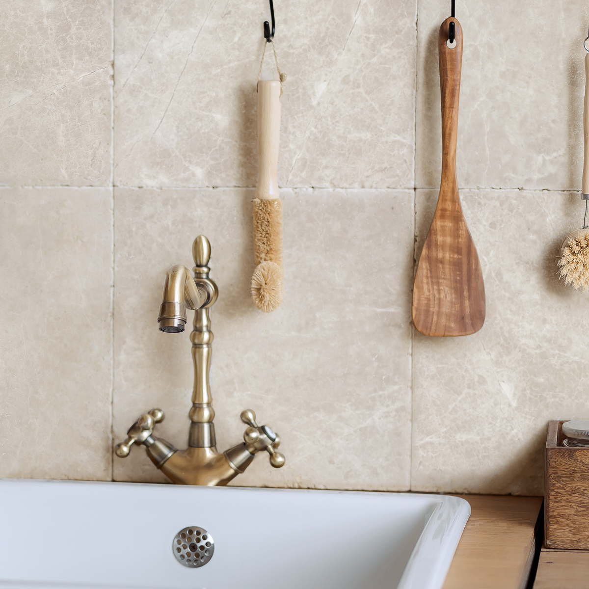 A kitchen scene with a white sink, a brass cross-handle faucet, cream marble wall tiles creating a splashback, and hanging wooden utensils.