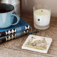 A square marble coaster with dalmatian dogs playing next to an old stone wall. The coaster is on a bedroom table next to books and a mug.
