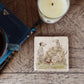 A square marble coaster with dalmatian dogs playing next to an old stone wall. The coaster is on a bedroom table next to books and a mug.