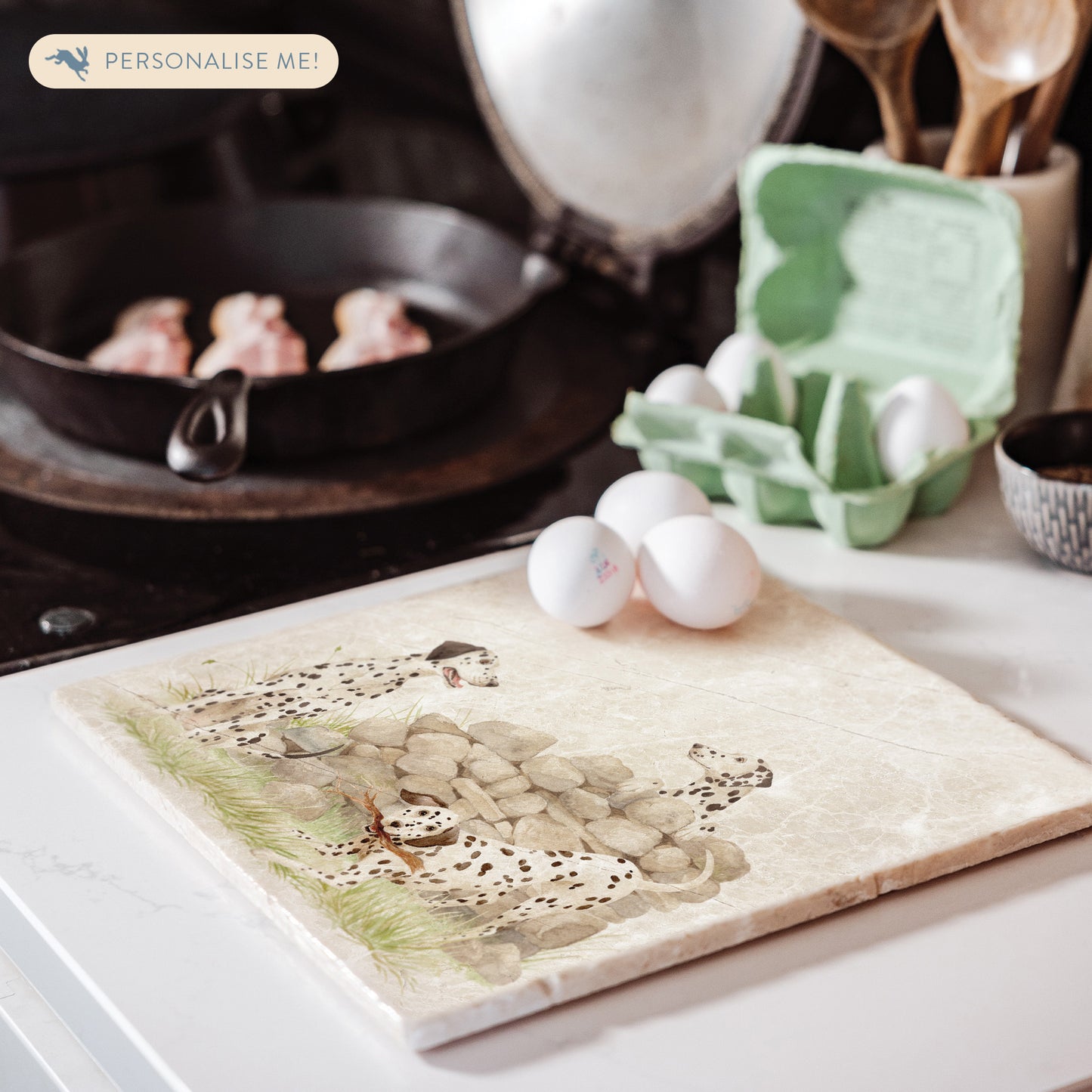 A large square marble platter featuring a  watercolour design of 3 dalmatian dogs playing fetch in the countryside. The platter is sat next to a stove top in the kitchen.