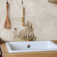A decorative kitchen backsplash tile featuring a Dalmatian dog illustration above a white sink and brass faucet, with hanging wooden kitchen tools on a beige tiled wall.