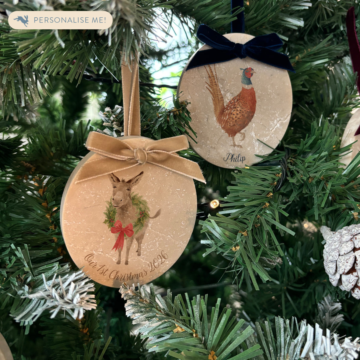 Two oval marble Christmas tree ornaments hanging on a Christmas tree. One ornament features a donkey wearing a green wreath with a red bow, with the text 'Our 1st Christmas' and the other features a pheasant with a personalised name underneath.