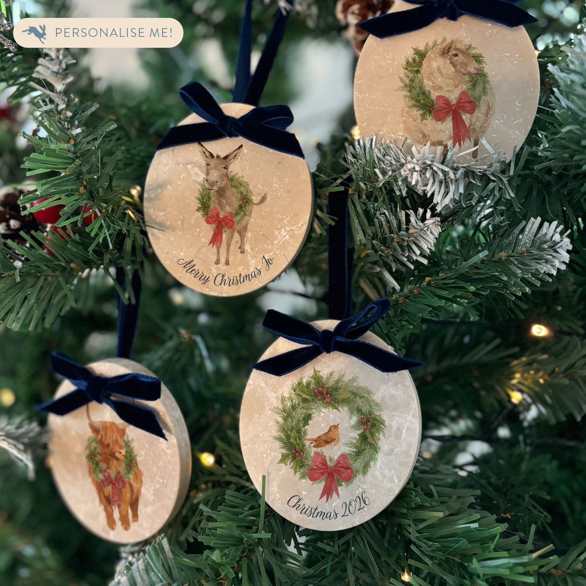 Four oval marble Christmas tree ornaments hanging on a Christmas tree. The baubles feature a donkey, highland cow, sheep, and robin all with festive wreaths. Some of the decorations are personalised with names and phrases.