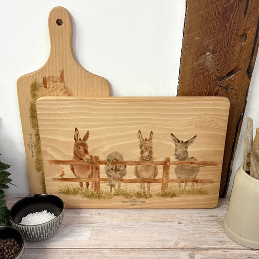 Rectangle wooden chopping board on a kitchen worktop. The beech wood board features  a watercolour design of cheeky donkeys peering over their fence.