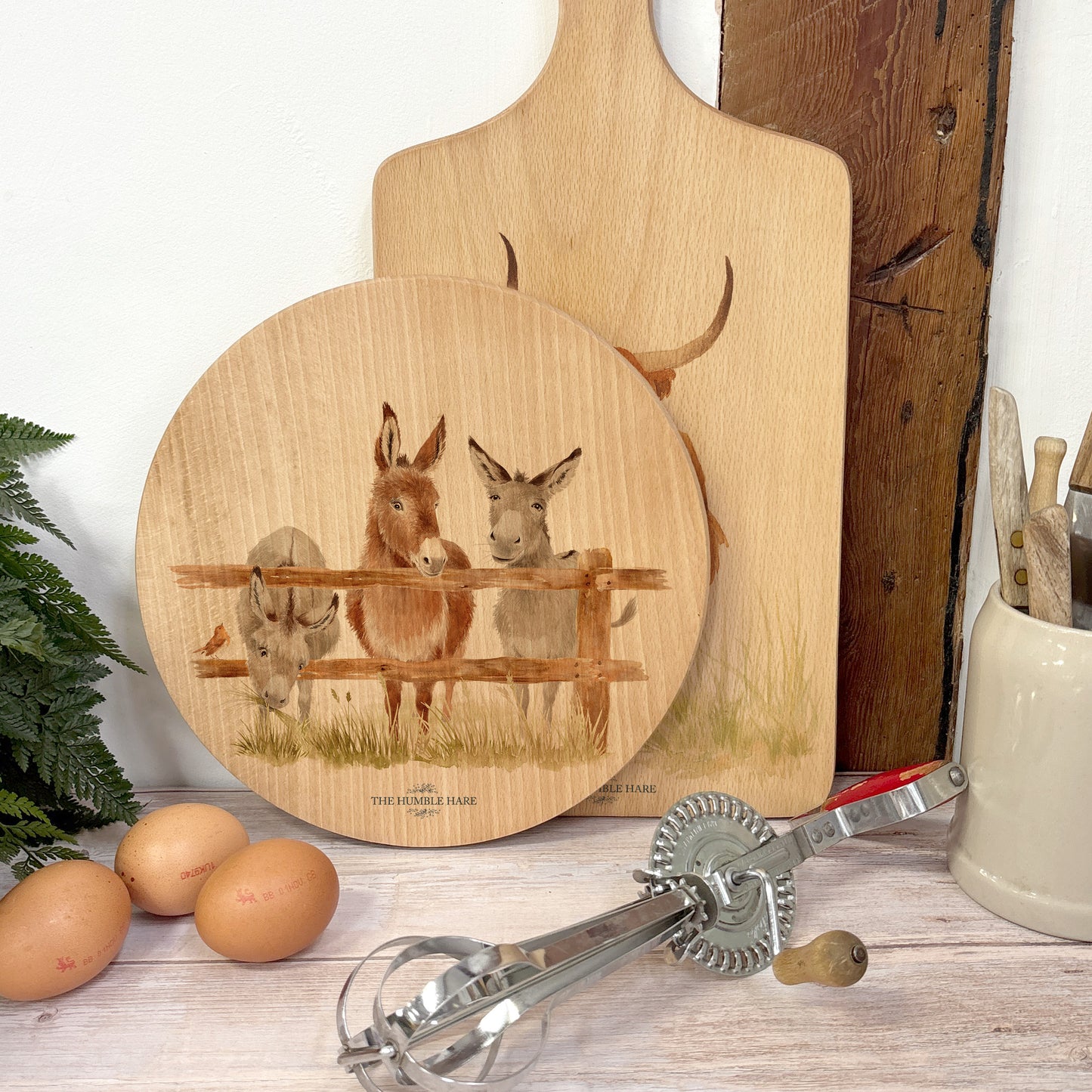 Round wooden chopping board on a kitchen worktop. The beech wood board features a watercolour design of cheeky donkeys peering over their fence.