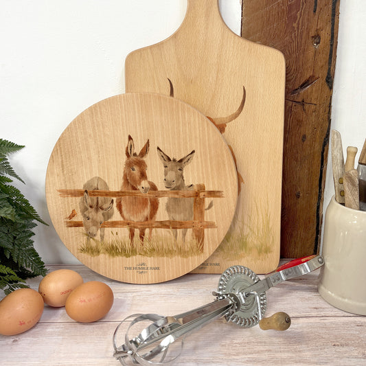 Round wooden chopping board on a kitchen worktop. The beech wood board features a watercolour design of cheeky donkeys peering over their fence.