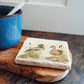 A handmade marble coaster featuring a watercolour design of two mallard ducks standing at the edge of a pond. The coaster is set on a kitchen worksurface.