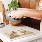 A large handmade marble platter being used as a trivet for a hot pan. The trivet features a watercolour design of two mallard ducks standing at the edge of a pond.