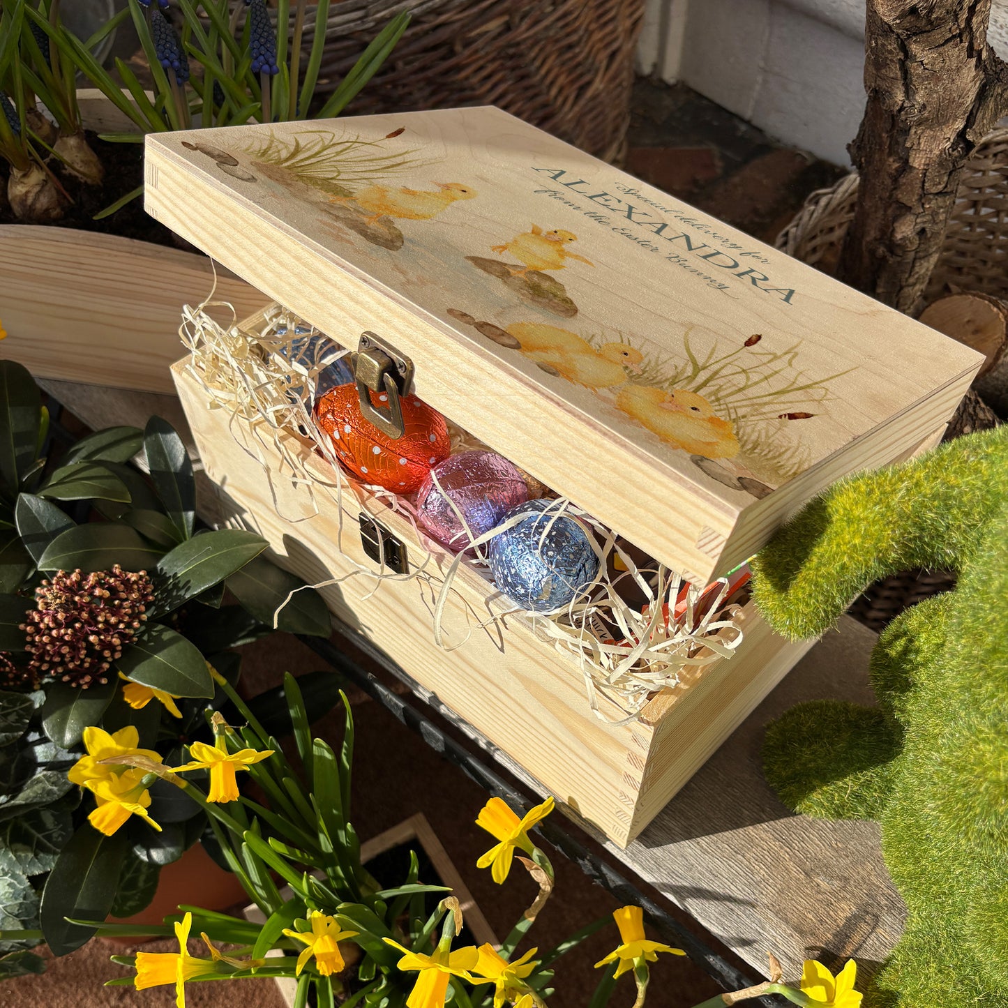 Small Easter personalised wooden pine box, perfect for Easter treats, eggs, gifting. Children gifts. This box has a beautiful easter theme with 4 fluffy ducklings playing in a pond area, surrounded by reeds and pebbles. This box can be personalised in 4 phrases or it can just have the design and no text. This box is showing an example of an easter box full with yummy easter treats, eggs, chocolate and biscuits. Perfect for gifting to parents, children and friends 