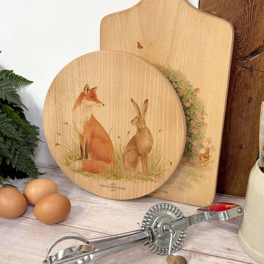 Round wooden chopping board on a kitchen worktop. The beech wood board features a watercolour design of a fox and hare in a meadow.