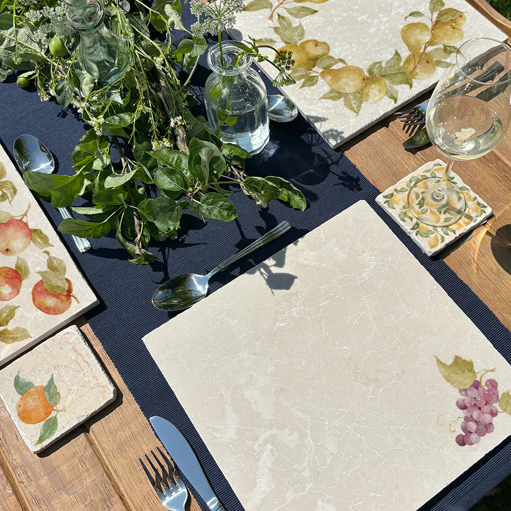 A farmhouse dining table set for a dinner party with marble placemats and coasters. The coasters and placemats have mix and match designs featuring different fruits.